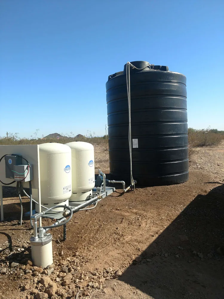 A black water storage tank with two pressure tanks and a wellhead on a desert lot